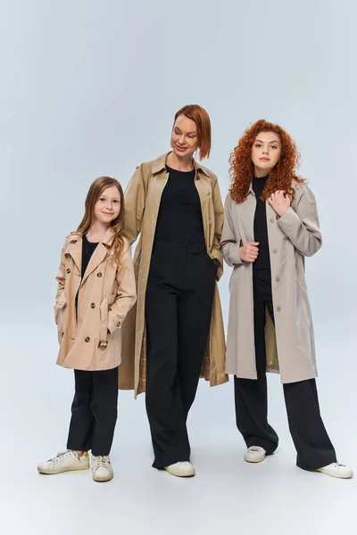 Famille rousse en trench coats posant ensemble sur fond gris, trois générations de femmes — Photo de stock