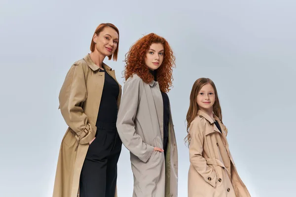 Happy redhead family in coats posing with hands in pockets on grey background, female generations — Stock Photo