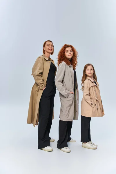 Alegre familia pelirroja en abrigos posando con las manos en bolsillos sobre fondo gris, generaciones femeninas - foto de stock
