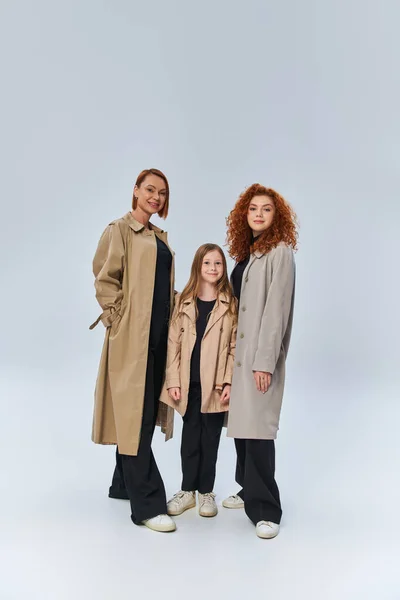 Familia pelirroja en abrigos de otoño y de pie juntos sobre fondo gris, generaciones femeninas felices - foto de stock
