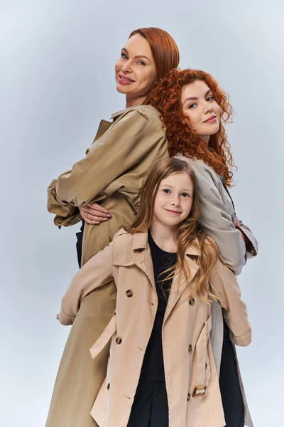 Cute redhead girl hugging happy relatives in autumn coats on grey background, female generations — Stock Photo