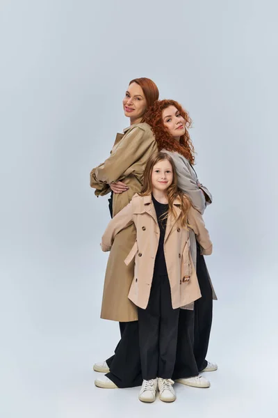 Rousse fille câlin heureux parents dans des manteaux sur fond gris, les générations féminines, pleine longueur — Photo de stock