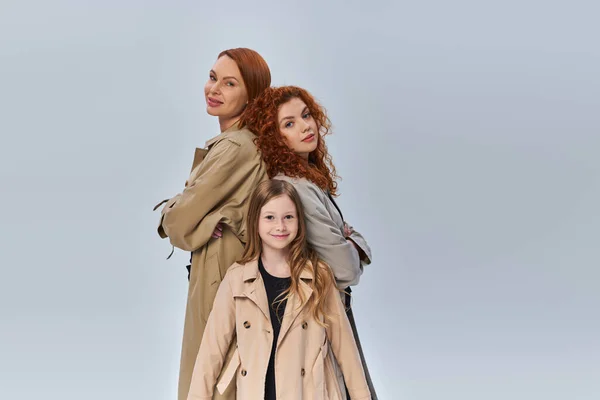 Redhead girl standing near happy relatives in coats on grey backdrop, female generation, fall style — Stock Photo