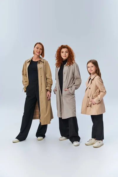 Руда сім'я в стильних пальто позує руками в кишенях на сірому тлі, три покоління — стокове фото