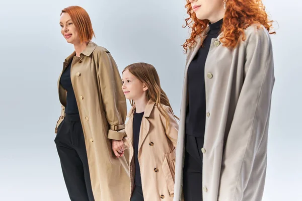 Familia pelirroja de tres generaciones caminando juntos en abrigos elegantes sobre fondo gris, moda de otoño - foto de stock