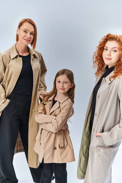 Три покоління радісної сім'ї, що стоїть разом в осінніх пальто на сірому фоні, осінь мода — стокове фото