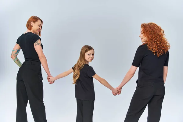 Three generations, happy girl holding hands with women, standing in matching attire on grey backdrop — Stock Photo