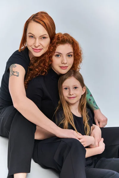Концепція трьох поколінь, радісна руда сім'я у відповідних нарядах, обіймаючись на сірому тлі — стокове фото