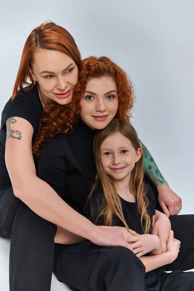 Concetto di tre generazioni, famiglia rossa positiva in abiti coordinati che si abbracciano su sfondo grigio — Foto stock