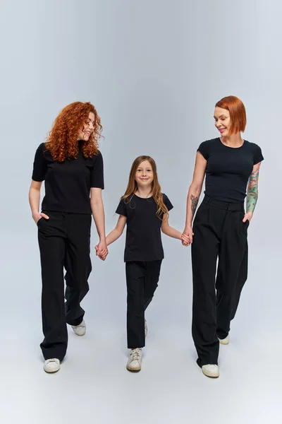 Fröhliches Mädchen hält Händchen und spaziert mit rothaariger Familie in passenden Outfits vor grauem Hintergrund — Stockfoto