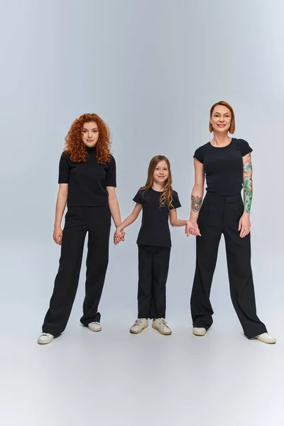 Cheerful girl holding hands and standing with redhead women in matching outfits on grey backdrop — Stock Photo