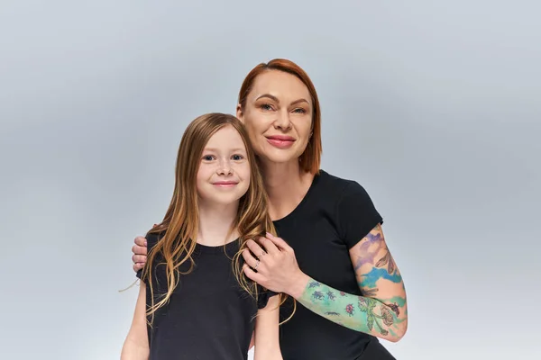Happy woman with tattoo hugging redhead girl and looking at camera on grey backdrop, generations — Stock Photo