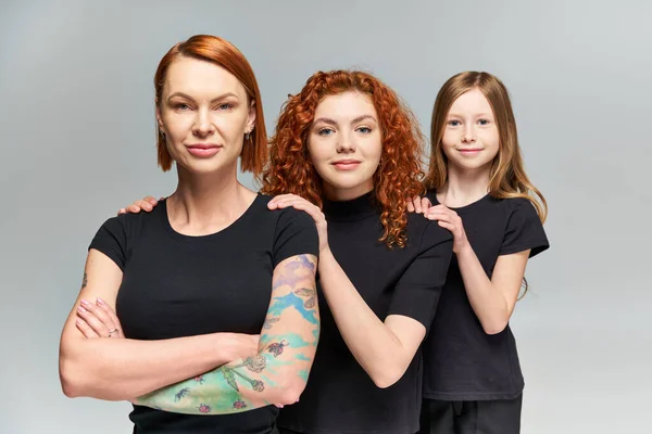 Three generations of happy women and girl posing in matching outfits on grey backdrop, family — Stock Photo