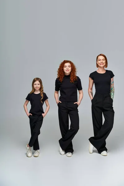 Tres generaciones de mujeres posando con las manos en los bolsillos en traje a juego sobre fondo gris - foto de stock