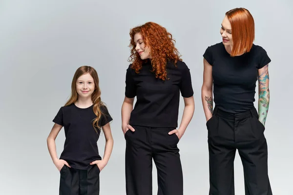 Tres generaciones, mujeres y niñas posando con las manos en los bolsillos en traje a juego sobre fondo gris - foto de stock