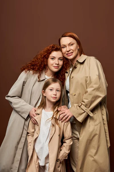 Three generations, cheerful women and girl posing in autumn trench coats on grey background, fashion — Stock Photo