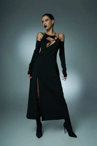 Espeluznante glamour, seductora mujer con labios oscuros posando en vestido de halloween negro en gris, longitud completa — Stock Photo