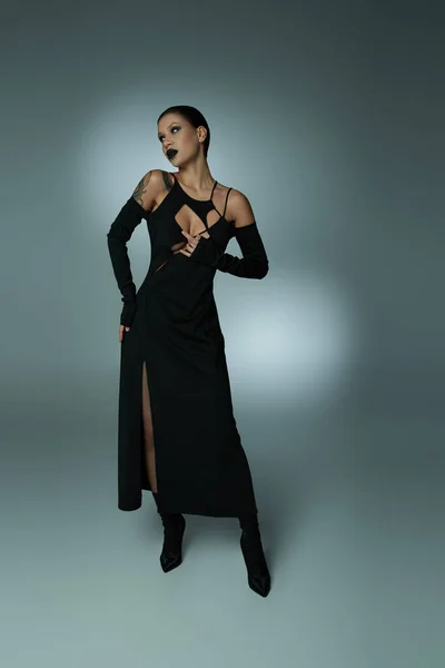 Belleza siniestra, mujer con maquillaje espeluznante oscuro posando en vestido de halloween negro en gris, longitud completa - foto de stock