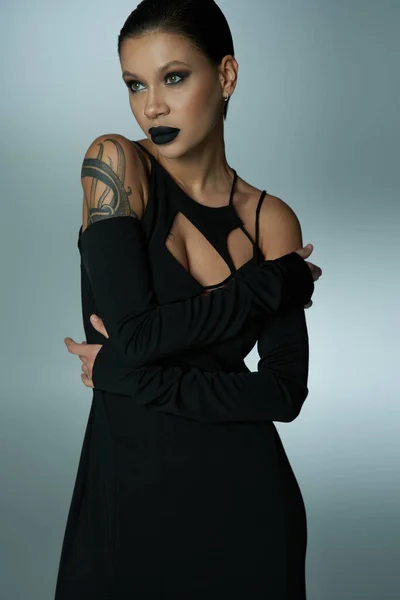 Misteriosa mujer tatuada con maquillaje de hechicera posando en vestido negro en gris, concepto de halloween - foto de stock