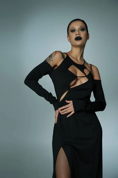 Mujer tatuada con maquillaje oscuro espeluznante vestido de halloween negro y mirando a la cámara en gris - foto de stock