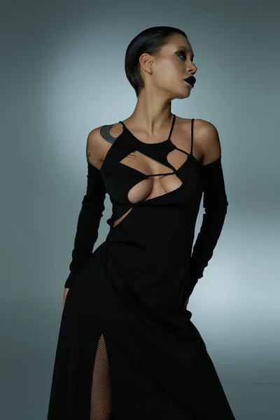 Concepto de Halloween, mujer sexy en el maquillaje de la hechicera y vestido elegante negro mirando hacia otro lado en gris - foto de stock