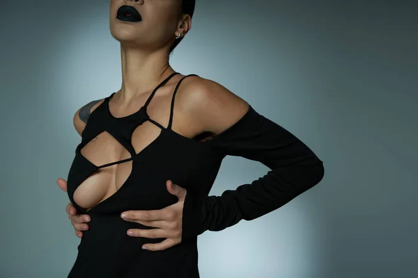 Femme tatouée sexy avec des lèvres foncées et les mains sous le sein posant en robe noire d'Halloween sur gris — Photo de stock