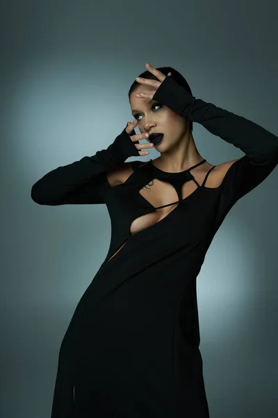 Belleza diabólica, mujer con estilo en el maquillaje misterioso y traje de halloween negro mirando hacia otro lado en gris - foto de stock