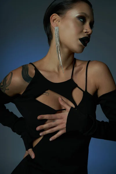 Mujer tatuada en vestido elegante negro y maquillaje siniestro mirando hacia otro lado en el fondo azul y gris - foto de stock