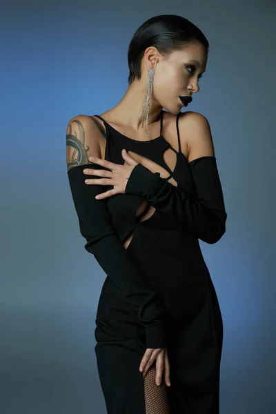 Готический гламур, татуированная женщина в черном платье и темный макияж глядя в сторону на синий анг серый фон — стоковое фото