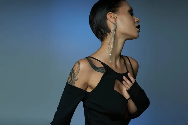 Mujer tatuada en vestido elegante negro y maquillaje oscuro sobre fondo azul y gris, belleza mágica - foto de stock
