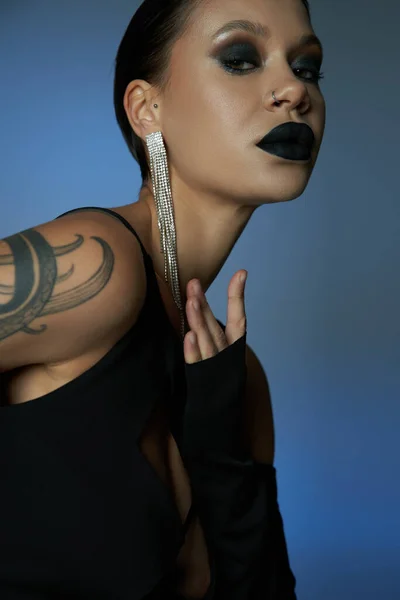 Retrato de mujer encantadora tatuada con maquillaje oscuro mirando a la cámara en el fondo azul y gris - foto de stock