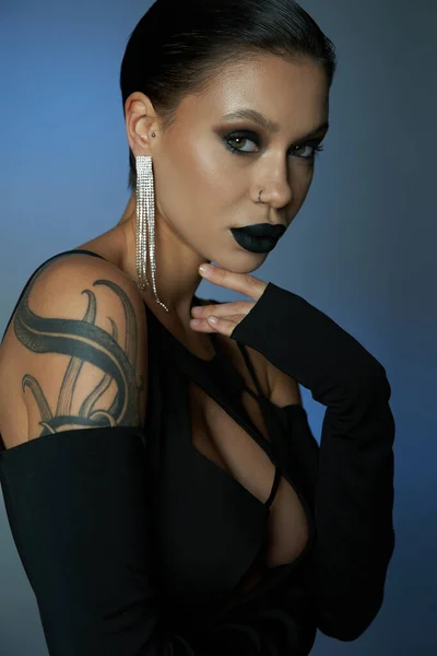 Mujer tatuada con maquillaje misterioso oscuro mirando a la cámara en el fondo azul y gris, concepto de Halloween - foto de stock