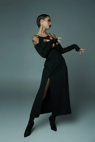 Темная красота, татуированная женщина в черном платье на Хэллоуин и жуткий макияж, позирующий на сером, полной длины — стоковое фото
