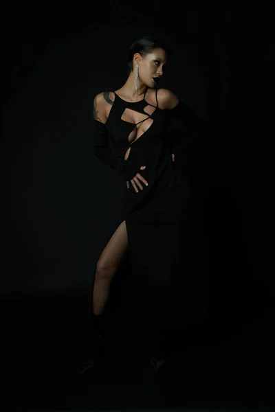 Волшебная чувственность, татуированная женщина в черном сексуальном платье позирует в темноте в студии, концепция Хэллоуина — стоковое фото
