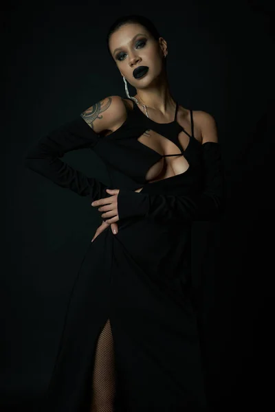 Tattooed woman in dark makeup and sexy halloween dress looking at camera in dark studio, black magic — Stock Photo