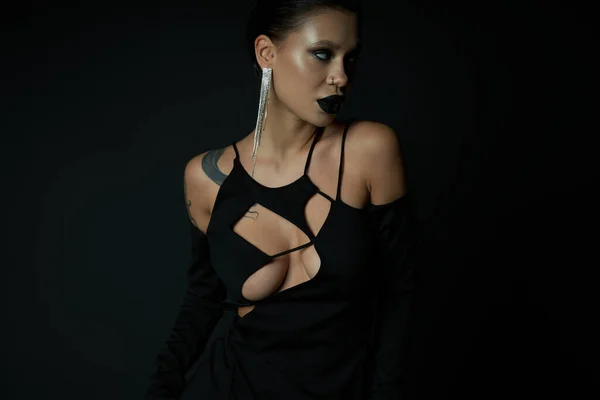 Mujer tatuada en pendiente brillante y vestido de bruja oscura mirando hacia otro lado en negro, concepto de halloween - foto de stock