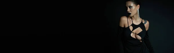Femme tatouée en boucle d'oreille brillante et robe de sorcière sombre sur fond noir, concept halloween, bannière — Photo de stock