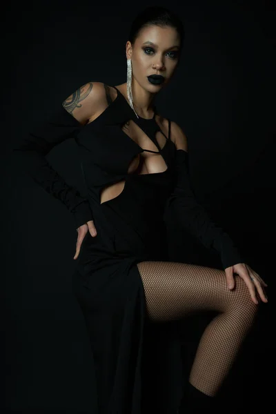 Stylische tätowierte Frau in schwarzem Halloween-Kleid und gruseligem Make-up posiert mit der Hand an der Hüfte auf schwarz — Stockfoto