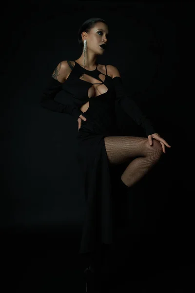 Seductora mujer tatuada en vestido de halloween negro y maquillaje oscuro mirando hacia otro lado mientras posa en negro - foto de stock