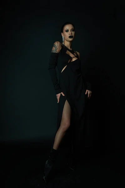 Verführerische tätowierte Frau in schwarzem Halloween-Kleid und dunklem Make-up, die wegschaut, während sie auf schwarz posiert — Stockfoto