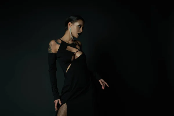 Gothic sensuality, tattooed woman in sexy enchantress dress and dark makeup looking away on black — Stock Photo
