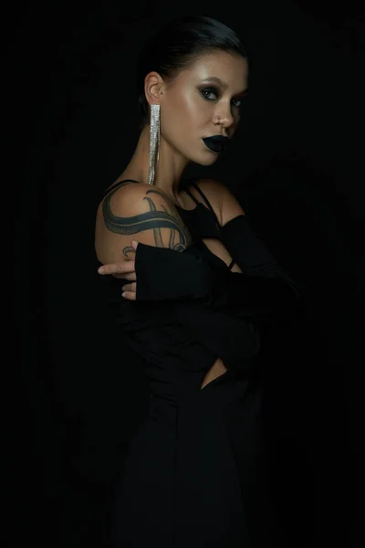 Соблазнительная татуированная женщина в блестящей сережке и ведьмином макияже, смотрящая в камеру на черную, темную элегантность — стоковое фото