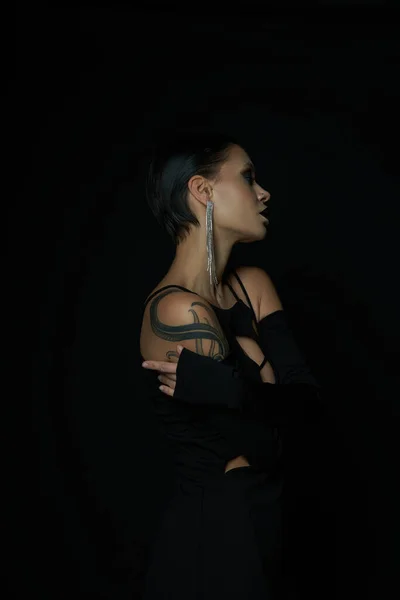 Side view of enchanting tattooed woman in black halloween dress and shiny earring on black backdrop — Stock Photo