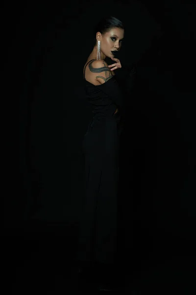 Таинственная татуированная женщина с темным макияжем и блестящей сережкой, смотрящая на камеру на черном фоне — стоковое фото