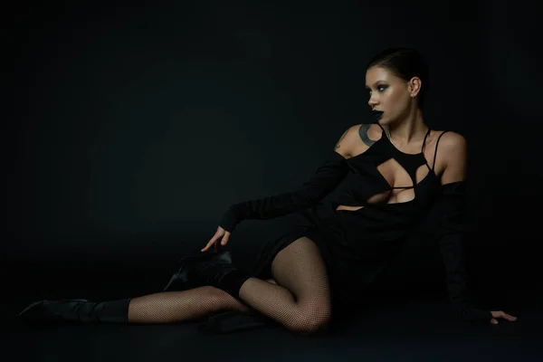 Mujer tatuada en traje de la encantadora oscura sentada y mirando hacia otro lado en negro, concepto de Halloween - foto de stock