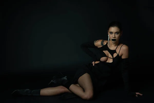 Sedutora mulher tatuada em vestido de Halloween sentado e olhando para a câmera em preto, sinistro beleza — Fotografia de Stock