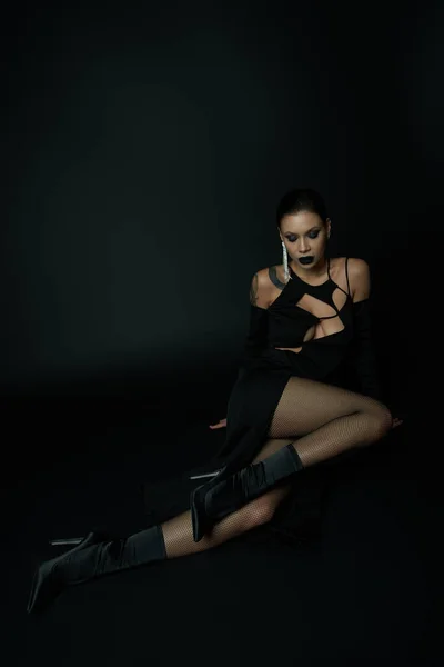 Hechizante mujer tatuada en vestido de halloween y maquillaje oscuro sentado en negro, glamour gótico - foto de stock