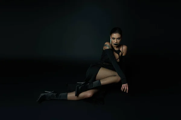 Stylische tätowierte Frau im Halloween-Kleid sitzt und schaut in die Kamera auf schwarzem, unheimlichem Glamour — Stockfoto
