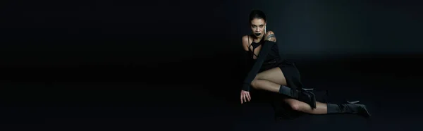 Sexy tatuado mulher no preto halloween vestido sentado no escuro e olhando para a câmera, banner — Fotografia de Stock
