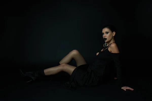 Mujer con estilo en traje de bruja oscura sentado y mirando a la cámara en negro, siniestra belleza - foto de stock